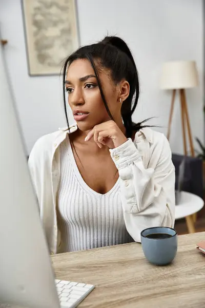 Eine junge schöne Frau studiert zu Hause, konzentriert und nippt an einem Getränk an ihrem Schreibtisch.. — Stockfoto