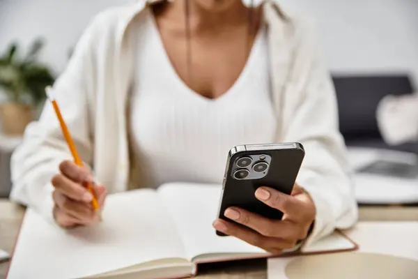 The beautiful young woman focuses on her studies at home, utilizing her phone and notebook. — Stock Photo