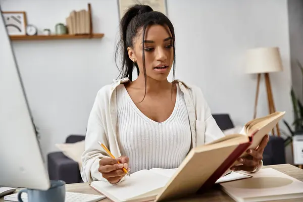 A focused young woman studies remotely, reading a book while taking notes at her cozy home desk. — Stock Photo