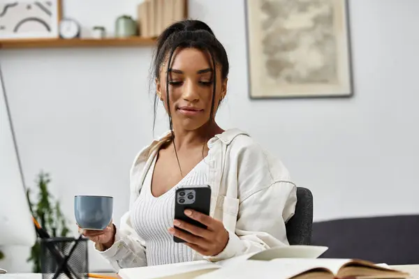 Una donna concentrata studia alla scrivania, sorseggiando il suo drink e rivedendo il suo telefono per informazioni. — Foto stock