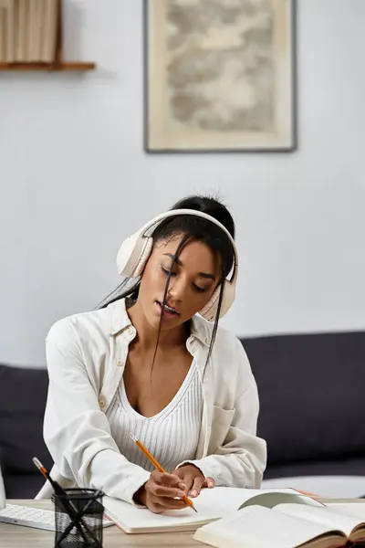 A young woman engages in remote studying at home, writing notes while wearing headphones. — Stock Photo