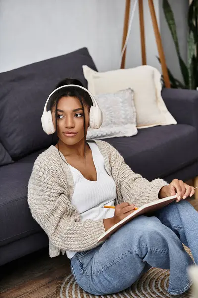 A young woman sits on the floor, taking notes in her notebook, while studying at home. — Stock Photo