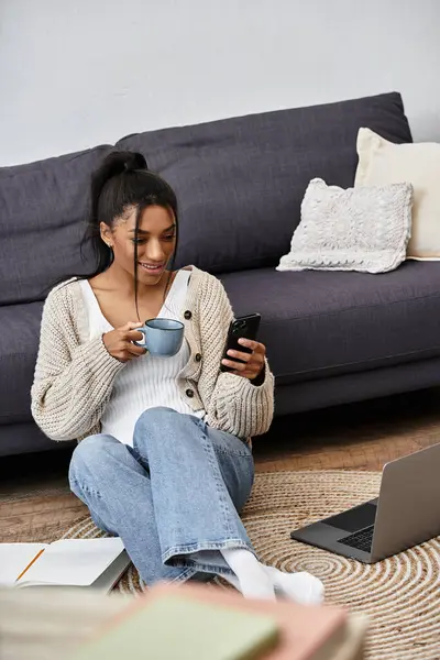 A cheerful woman studies from home, sipping tea and checking her phone while sitting comfortably. — Stock Photo