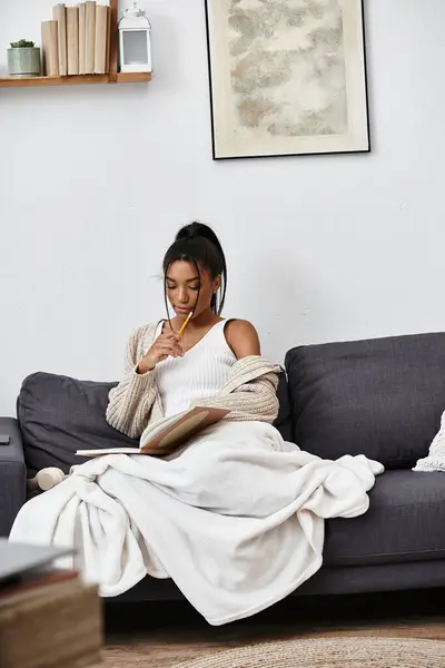 A young woman thoughtfully studies at home, wrapped in a cozy blanket while writing notes. — Stock Photo