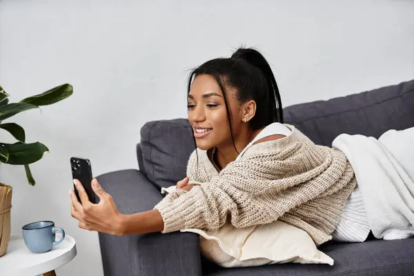 Una giovane donna studia attentamente da casa sua, sorridendo mentre interagisce con il suo smartphone. — Foto stock