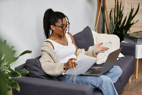 A young woman studies remotely, sitting on a couch surrounded by plants, looking focused. — Stock Photo