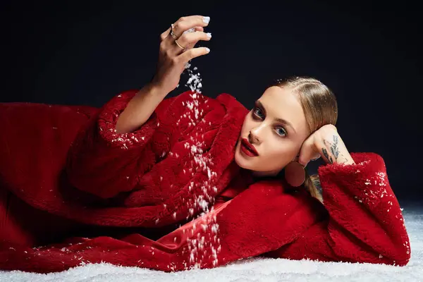 A young woman with an elegant demeanor sprinkles snow while lounging in a vibrant red coat. — Stock Photo