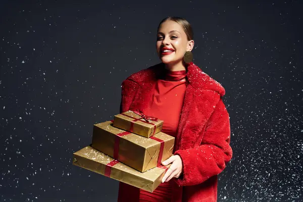 A stunning young woman in red holds beautifully wrapped gifts while enjoying a snowy celebration. — Stock Photo