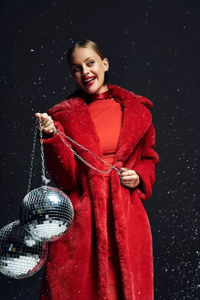 The young woman radiates joy, dressed in a striking red coat, holding shimmering disco balls. — стоковое фото