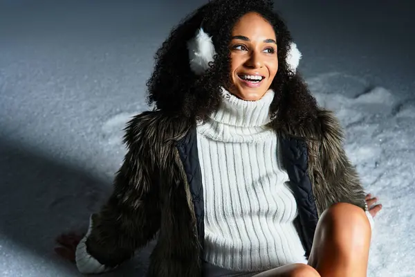 Una donna bruna elegante indossa paraorecchie e una giacca di pelliccia ecologica mentre posa in un ambiente da studio invernale. — Foto stock