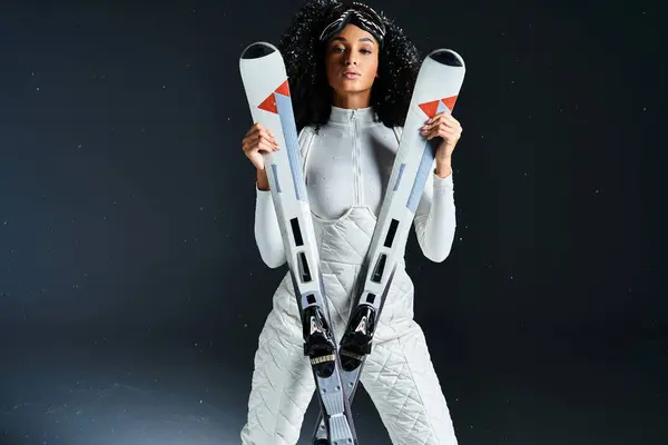 A stunning brunette woman dons a fashionable ski suit, holding skis in a winter-themed studio setting. — Stock Photo