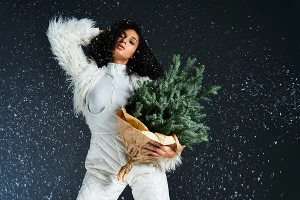 A stylish African American woman showcases winter fashion, holding pine branches while snowflakes fall around her. — Stock Photo