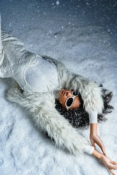 A stylish African American woman relaxes in the snow wearing a white ski ensemble and faux fur coat. — Stock Photo