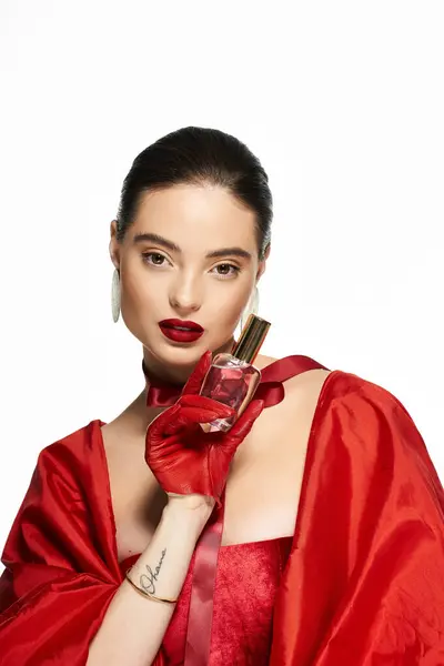 A stunning woman showcases a perfume bottle while dressed in luxurious red fabric. — Stock Photo