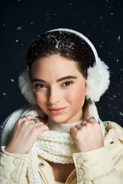 A woman smiles warmly while wearing ear muffs and a cozy scarf against falling snowflakes. — Stock Photo