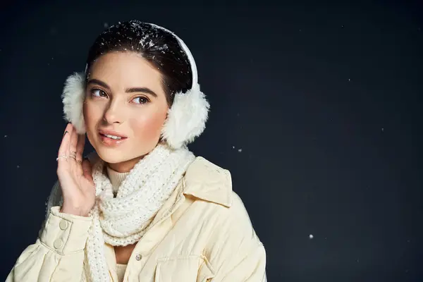 Una donna gioiosa che indossa paraorecchie e una sciarpa accogliente sorride mentre i fiocchi di neve cadono delicatamente intorno a lei.. — Foto stock
