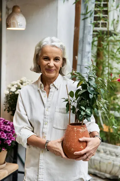 A mature woman smiles warmly while holding a lovely potted plant in a vibrant shop. — Stock Photo