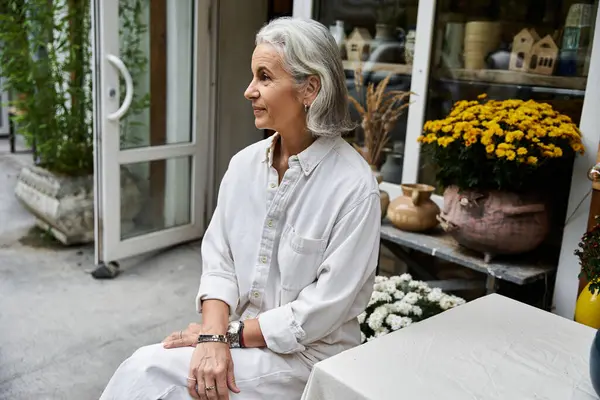 The elegant mature woman reflects in a charming outdoor setting filled with vibrant flowers. — Stockfoto