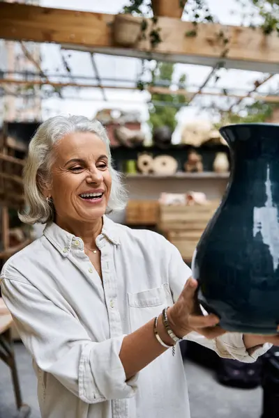 A mature woman smiles brightly while holding a stunning piece of pottery in her studio. — Stockfoto