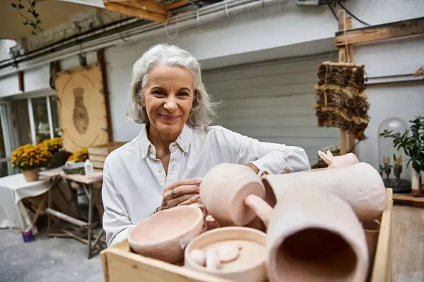 A talented mature woman smiles warmly while surrounded by her handmade pottery in a charming studio. — Stock Photo