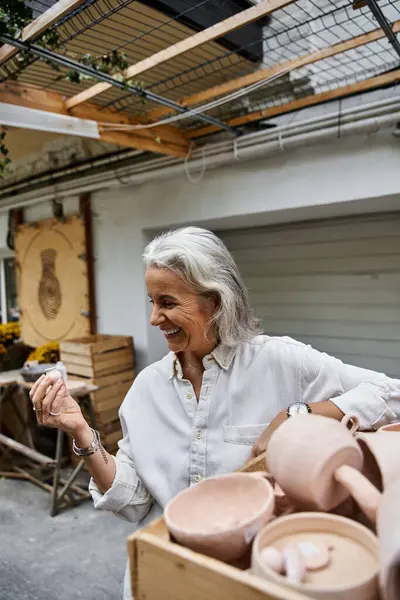 The mature woman smiles brightly while holding a piece of pottery in a vibrant workspace. — Stock Photo