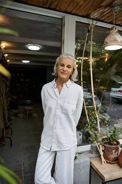 Una mujer madura con el pelo plateado sonríe calurosamente mientras disfruta del ambiente sereno de su tienda. - foto de stock