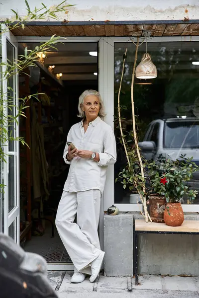 A sophisticated woman stands at the entrance of a boutique, lost in thought, holding her phone. — Stock Photo