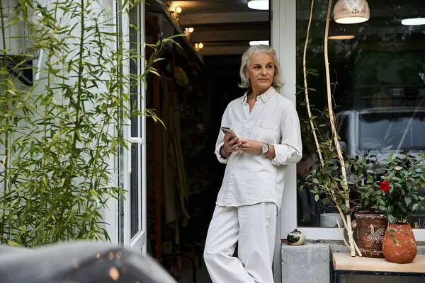 The graceful woman stands at the entrance of her cozy shop, engaged with her smartphone. — Stock Photo