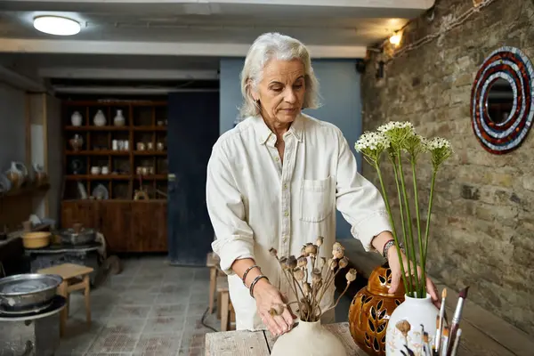 A beautiful mature woman carefully arranges delicate flowers in vases on a rustic wooden table. — Stock Photo