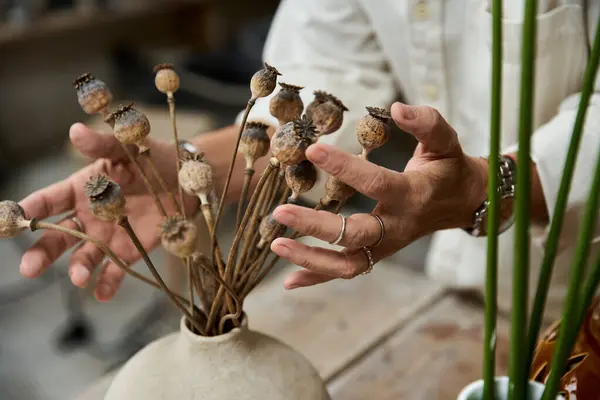 Una donna matura organizza delicatamente fiori di papavero secchi in un vaso rustico mentre si concentra intensamente.. — Foto stock