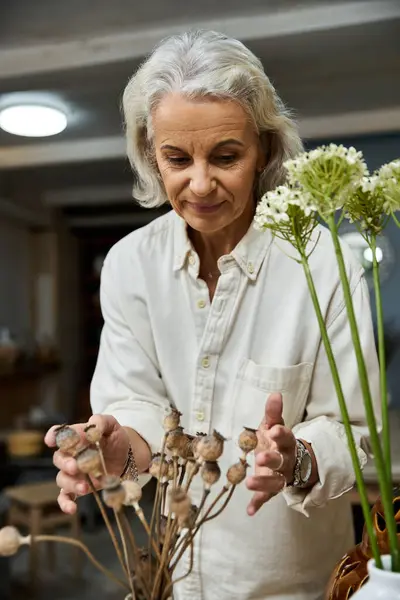 A mature woman artfully arranging dried floral stems in a serene, warmly lit space. — Stock Photo