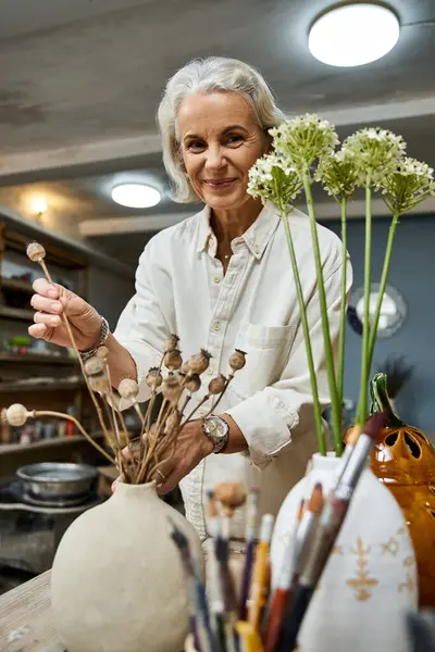 A graceful mature woman skillfully arranges dried flowers in a charming artistic setting. — Stock Photo