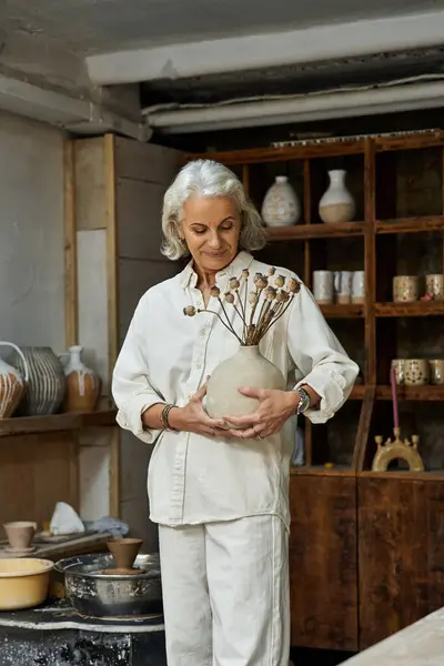The mature woman holds a unique vase filled with dried flowers, smiling softly in her studio. — Stock Photo