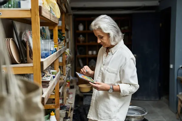 A graceful mature woman carefully paints in her artistic studio surrounded by handmade ceramics. — Stock Photo
