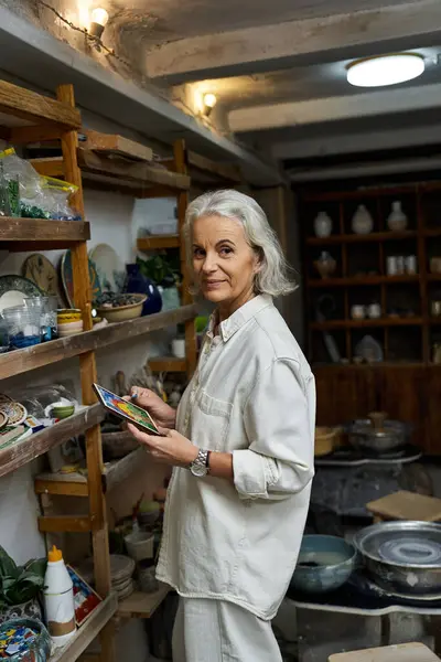 A beautiful mature woman examines her collection in a charming artistic studio filled with pottery. — Stock Photo