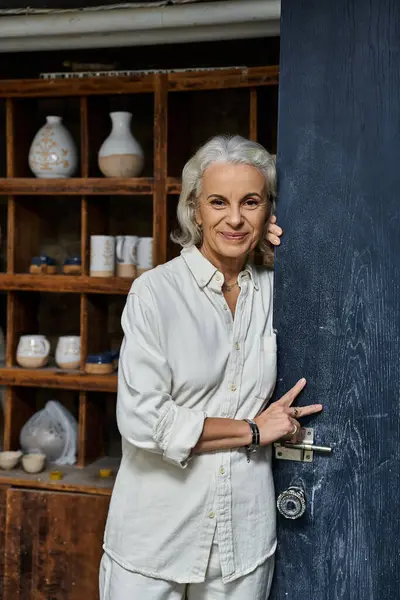 A cheerful woman stands at the doorway of her pottery studio, inviting creativity within. — Stock Photo