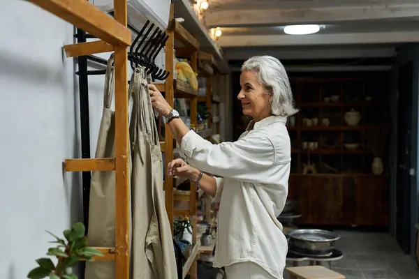 The mature woman gently arranges her garments on a wooden rack in a warmly lit space. — Stock Photo