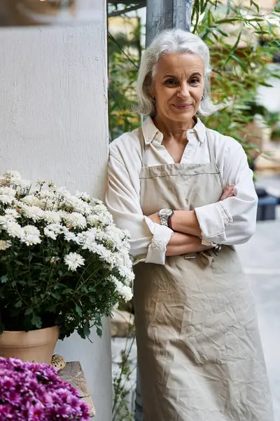 A mature woman in an apron smiles warmly while surrounded by vibrant flowers in a garden. — Stock Photo