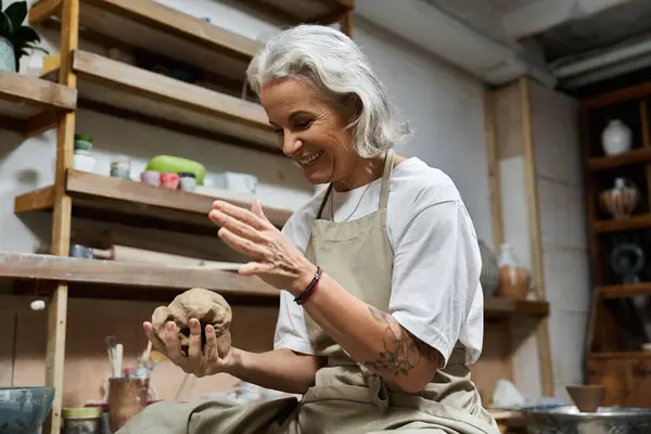 Eine reife Frau mit silbernem Haar lächelt, während sie in einem hellen, künstlerischen Arbeitsraum Ton formt. — Stockfoto