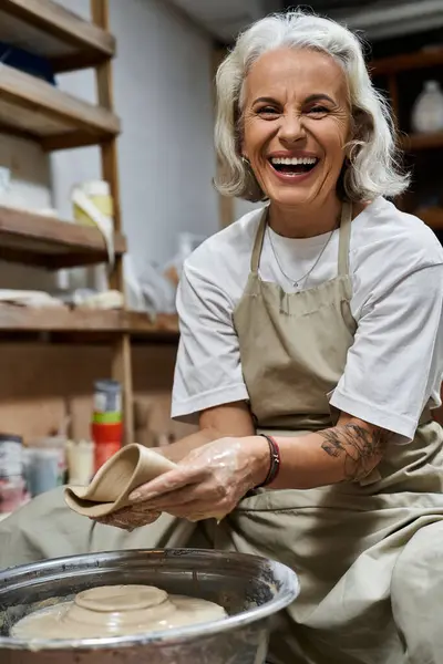 Eine reife Frau lächelt strahlend, während sie an ihrer Töpferscheibe Ton formt, umgeben von Kreativität. — Stockfoto