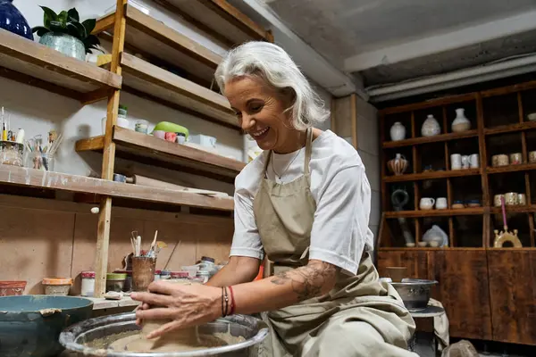A mature woman joyfully shapes clay in a cozy pottery studio filled with inspiration. — Stock Photo