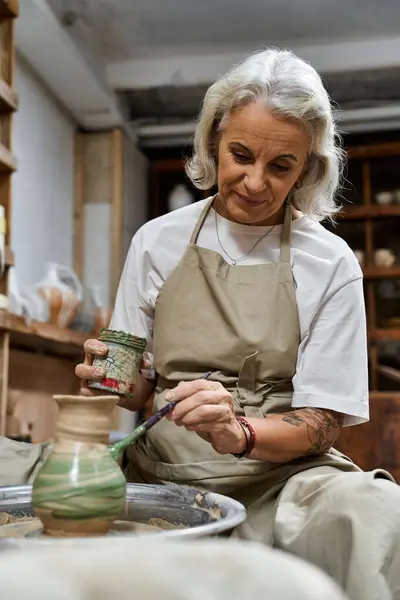 A mature woman adds intricate designs to a pottery item while sitting in her studio. — Stock Photo