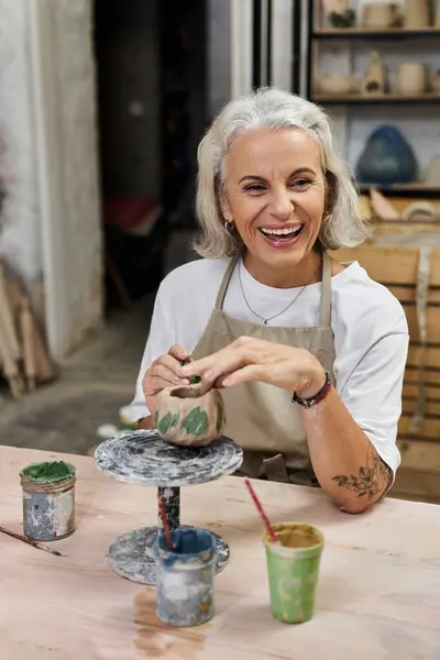 A mature woman smiles brightly while shaping clay, surrounded by colorful paint pots and tools. — Stock Photo
