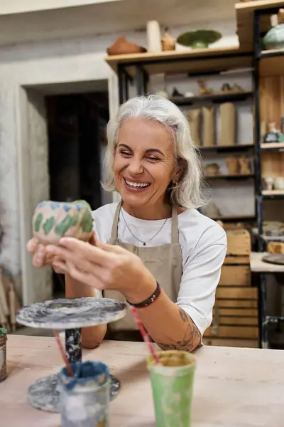 A mature woman smiles brightly while crafting a unique pottery piece in her artistic studio setting. — Stock Photo