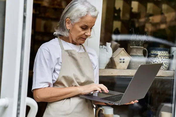 A beautiful mature woman in an apron engages with her laptop near a pottery shop filled with crafts. — Stock Photo