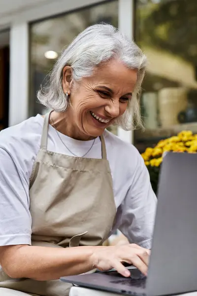 A cheerful woman smiles as she types on her laptop in a sunny garden filled with flowers. — Stock Photo