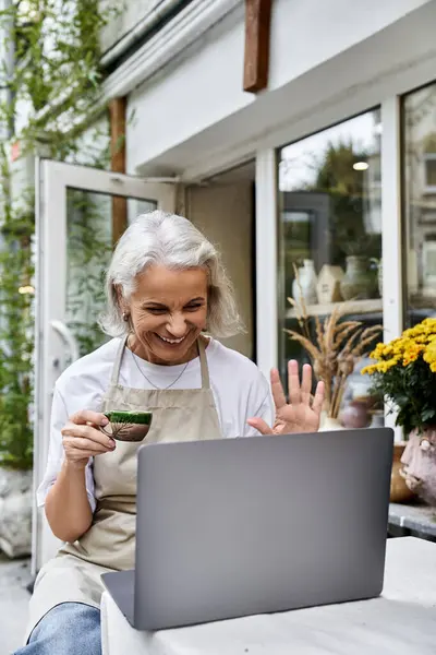 Uma mulher madura feliz saboreia café enquanto se envolve com alguém em seu laptop ao ar livre. — Fotografia de Stock