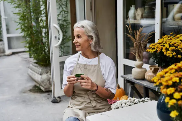 The mature woman holds a small object, surrounded by autumn flowers in a serene garden setting. — Stock Photo