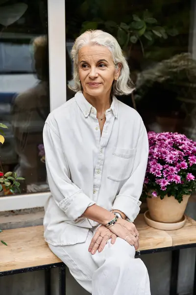 Une femme gracieuse aux cheveux argentés se détend joyeusement à côté de fleurs vibrantes dans un cadre paisible. — Photo de stock