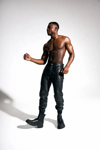 This striking portrait features a young, handsome African American man posing confidently without a shirt. He stands in stylish leather pants and bold boots, capturing an artistic and modern vibe. — Stock Photo